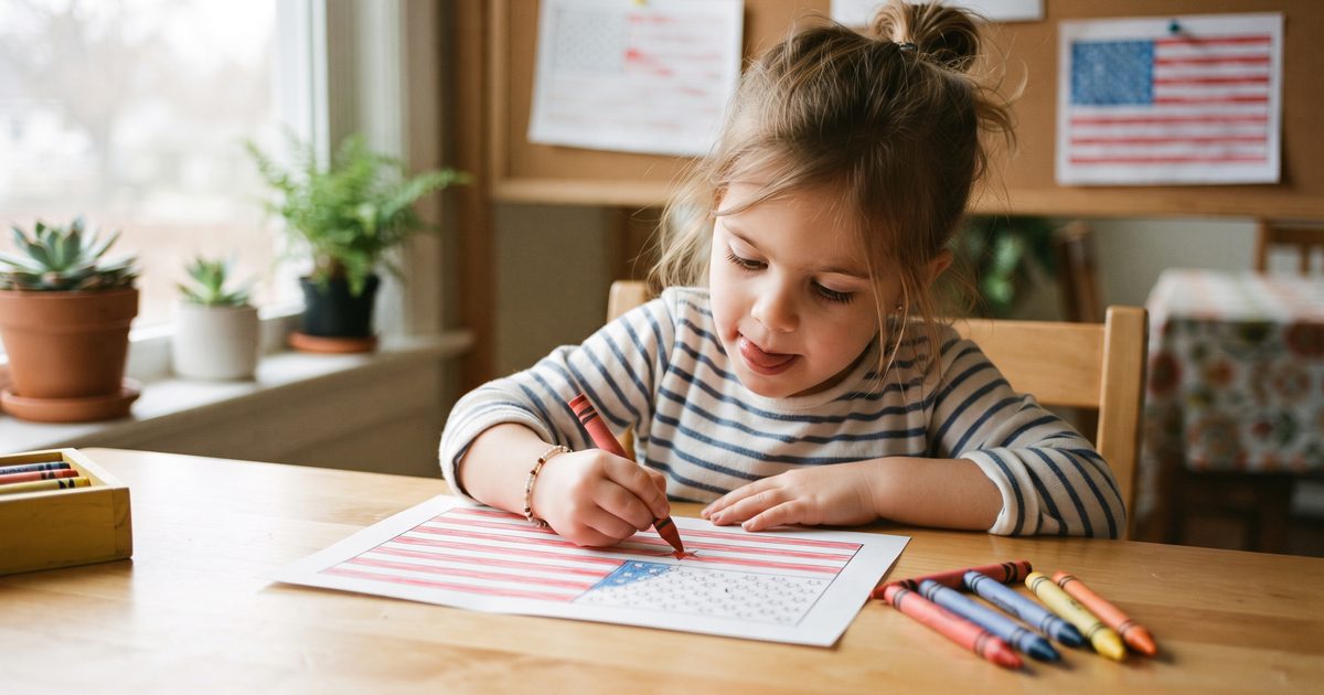 Zdjęcie z bliska przedstawiające dziewczynkę w wieku przedszkolnym, która czerwoną kredką starannie koloruje małą pięcioramienną gwiazdkę na kartce z flagą w kolorach czerwonym, białym i niebieskim, z językiem wysuniętym z koncentracji, obok garść zatemperowanych kredek i gotowa strona z flagą na drewnianym stole